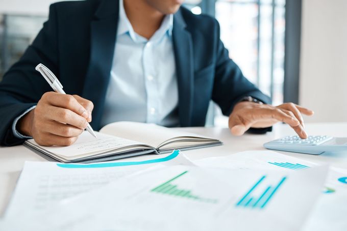 Person in a suit writing in a notebook at a desk with charts and financial documents.