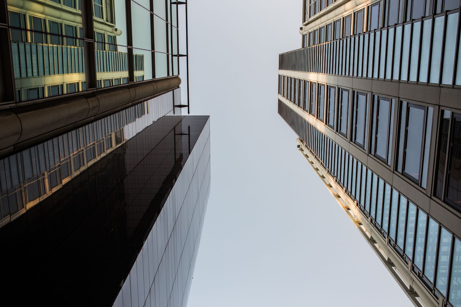 Looking up between two tall glass office buildings against a pale sky