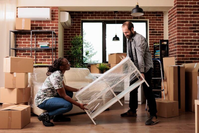 Two people unpacking furniture in a brick-walled apartment, with moving boxes and a couch nearby.