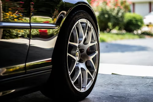 Close-up of a clean, black car's front wheel with a silver, multi-spoke rim parked on a concrete driveway.