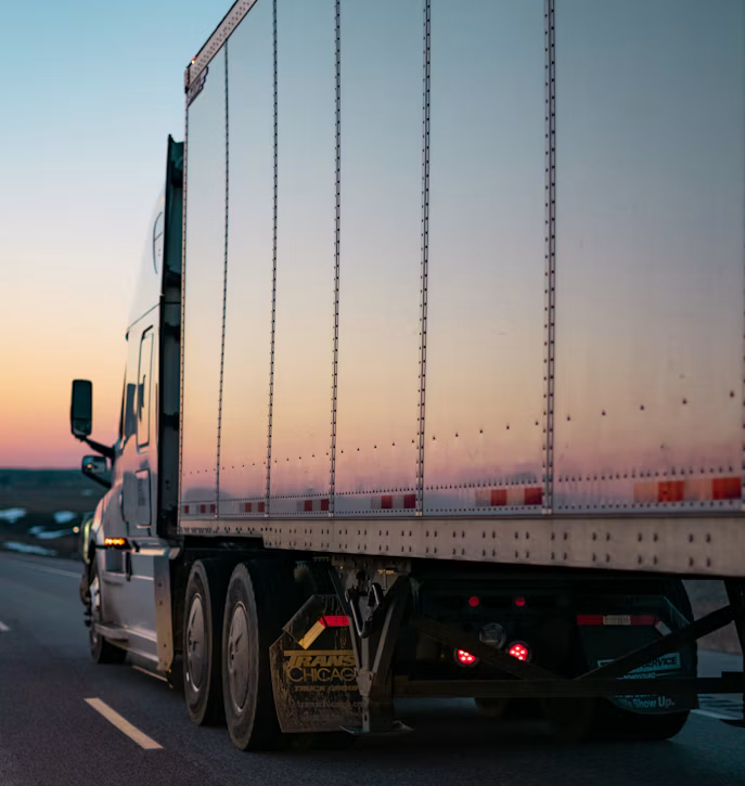 Semi-truck trailer driving on a road at sunset, with pink and blue sky reflections.