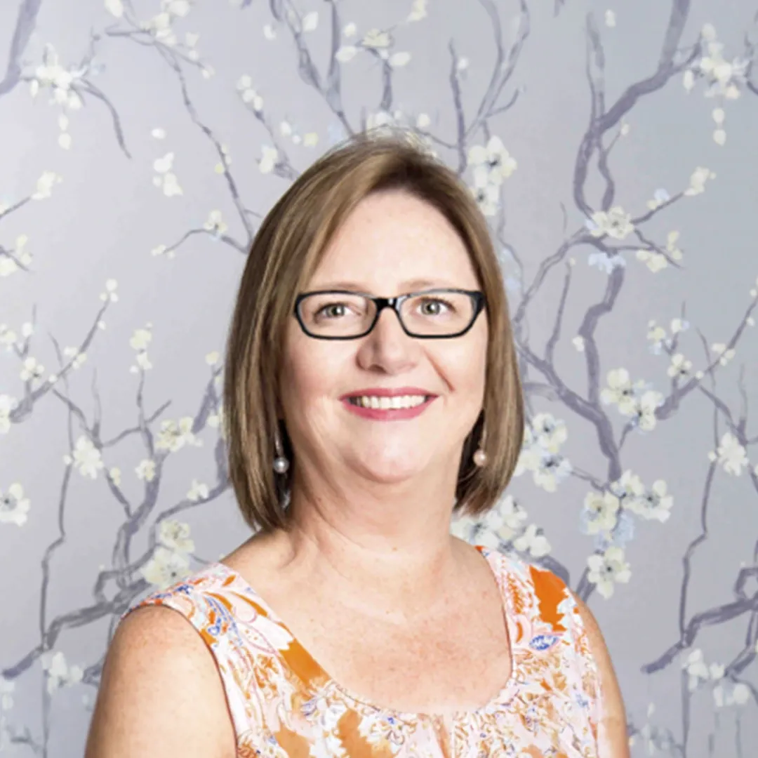 Portrait of a smiling woman with glasses in an orange floral sleeveless top against a gray floral backdrop