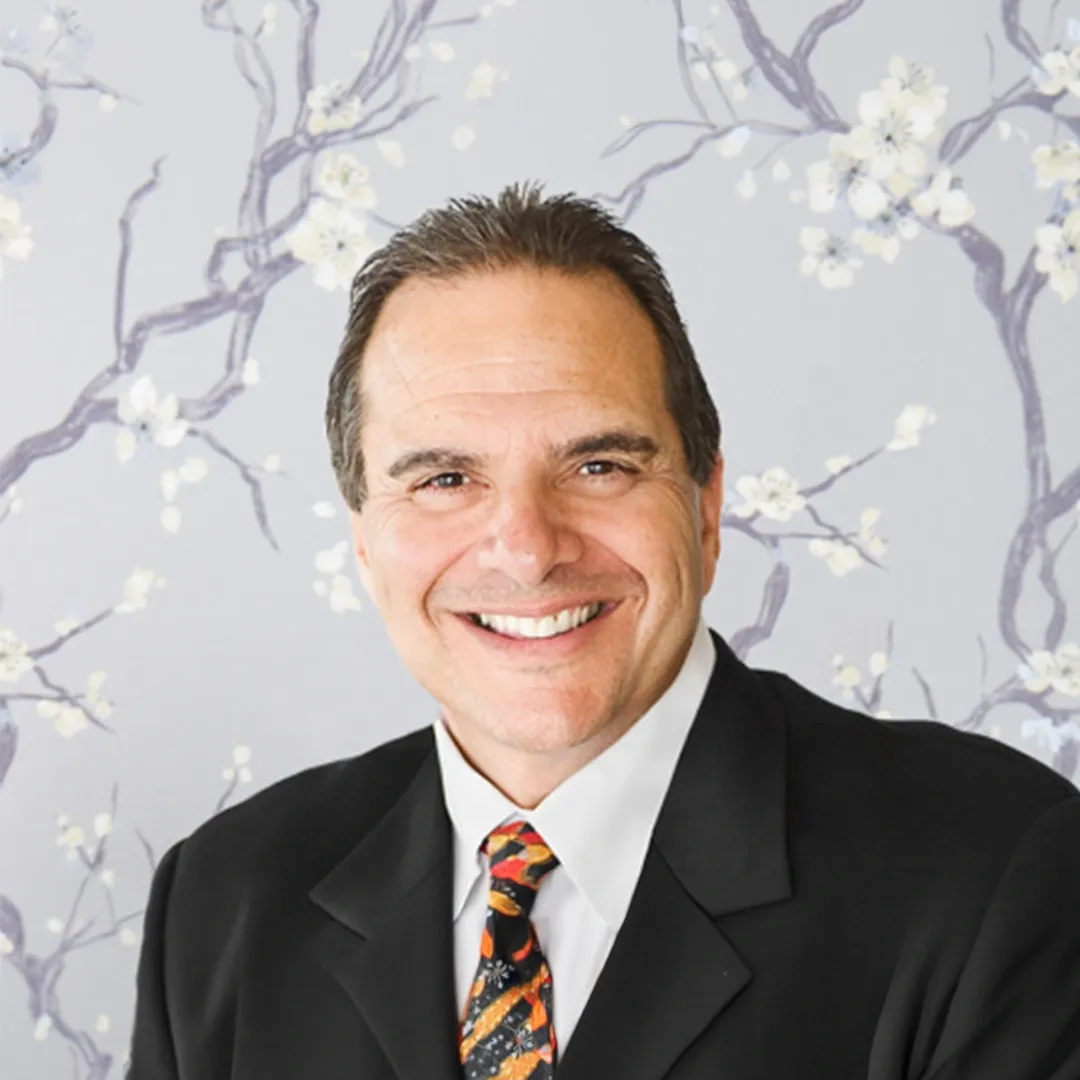 Smiling man in a black suit and patterned tie against a light floral background