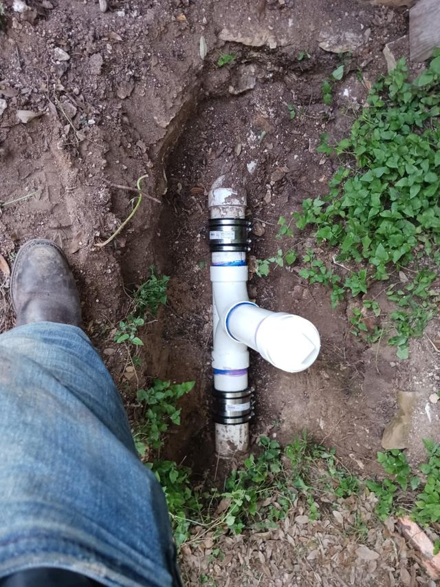 A buried white PVC pipe with a T-joint in a dirt trench next to a person’s boot and jeans.
