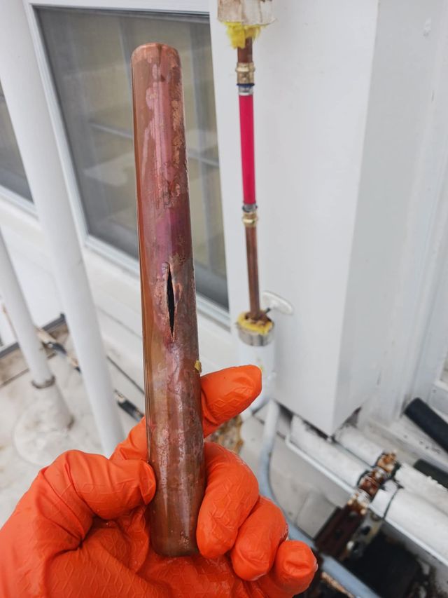 Gloved hand holding a rusted copper pipe in front of a house exterior and utility piping