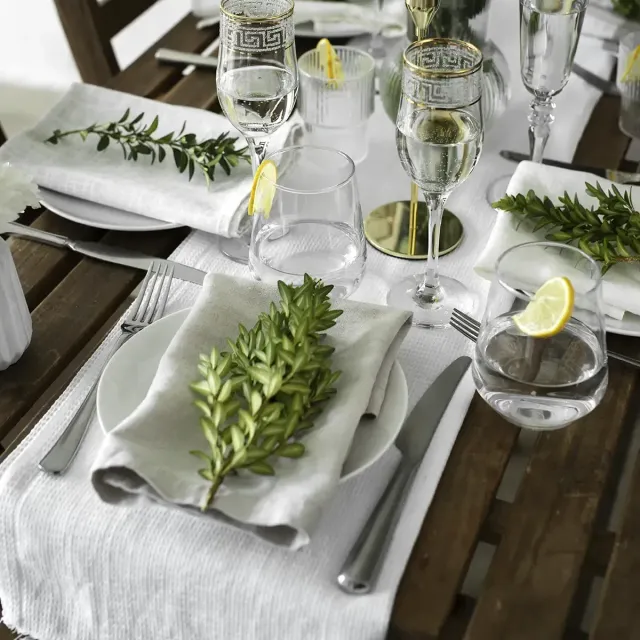 Elegant table setting with white dishes, green herbs, and lemon-garnished glassware on a wooden table.