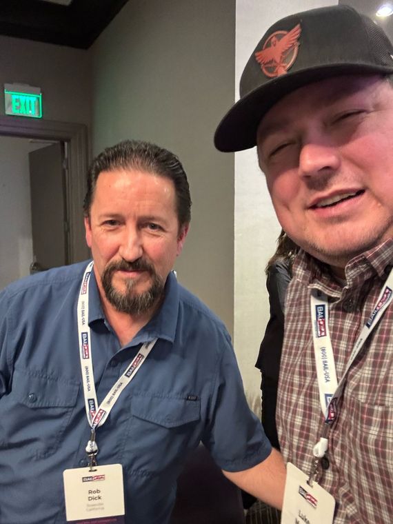 Two men wearing lanyards pose together indoors, one holding the camera in a hallway.