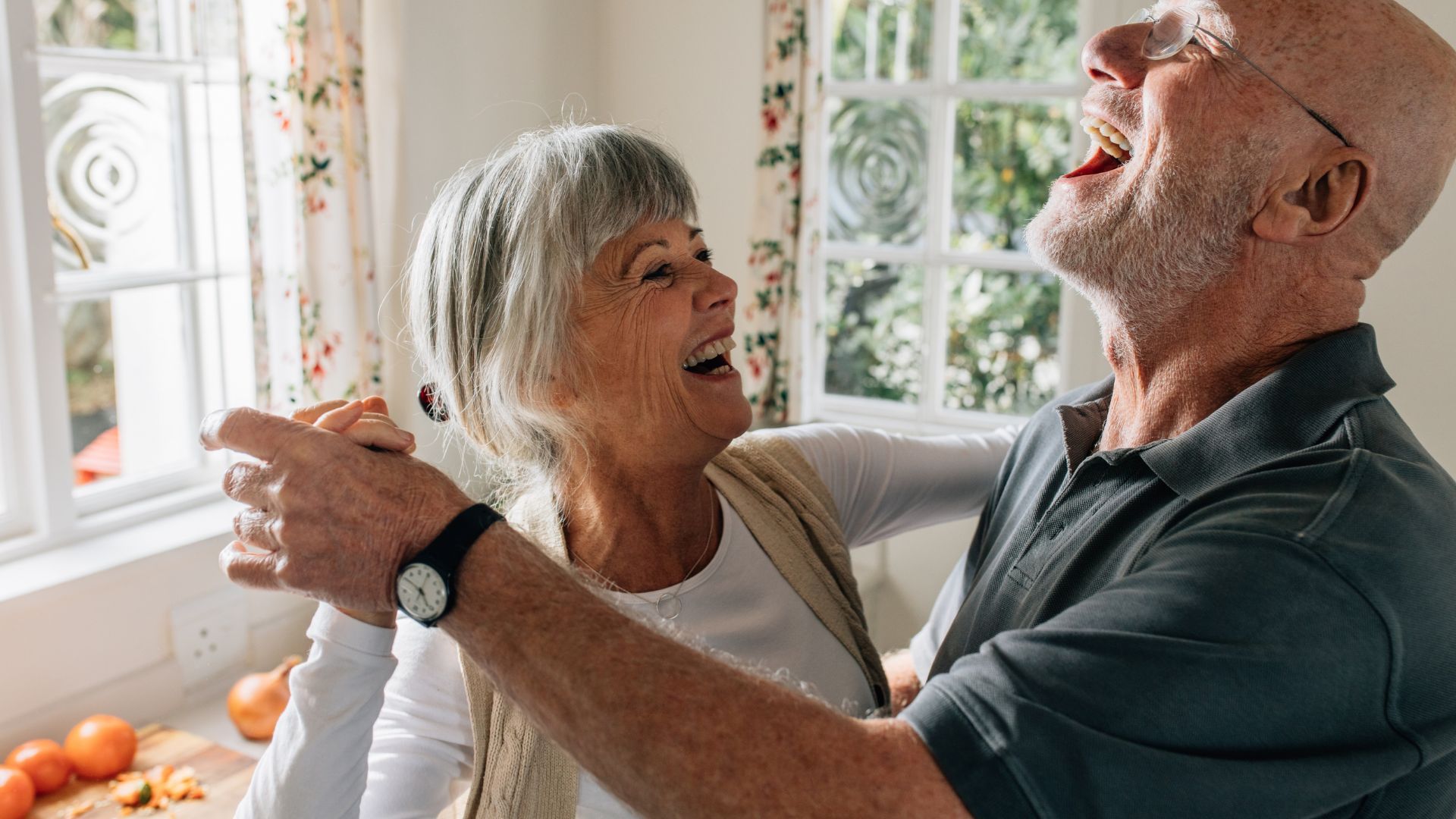 An elderly couple is dancing together in front of a window.