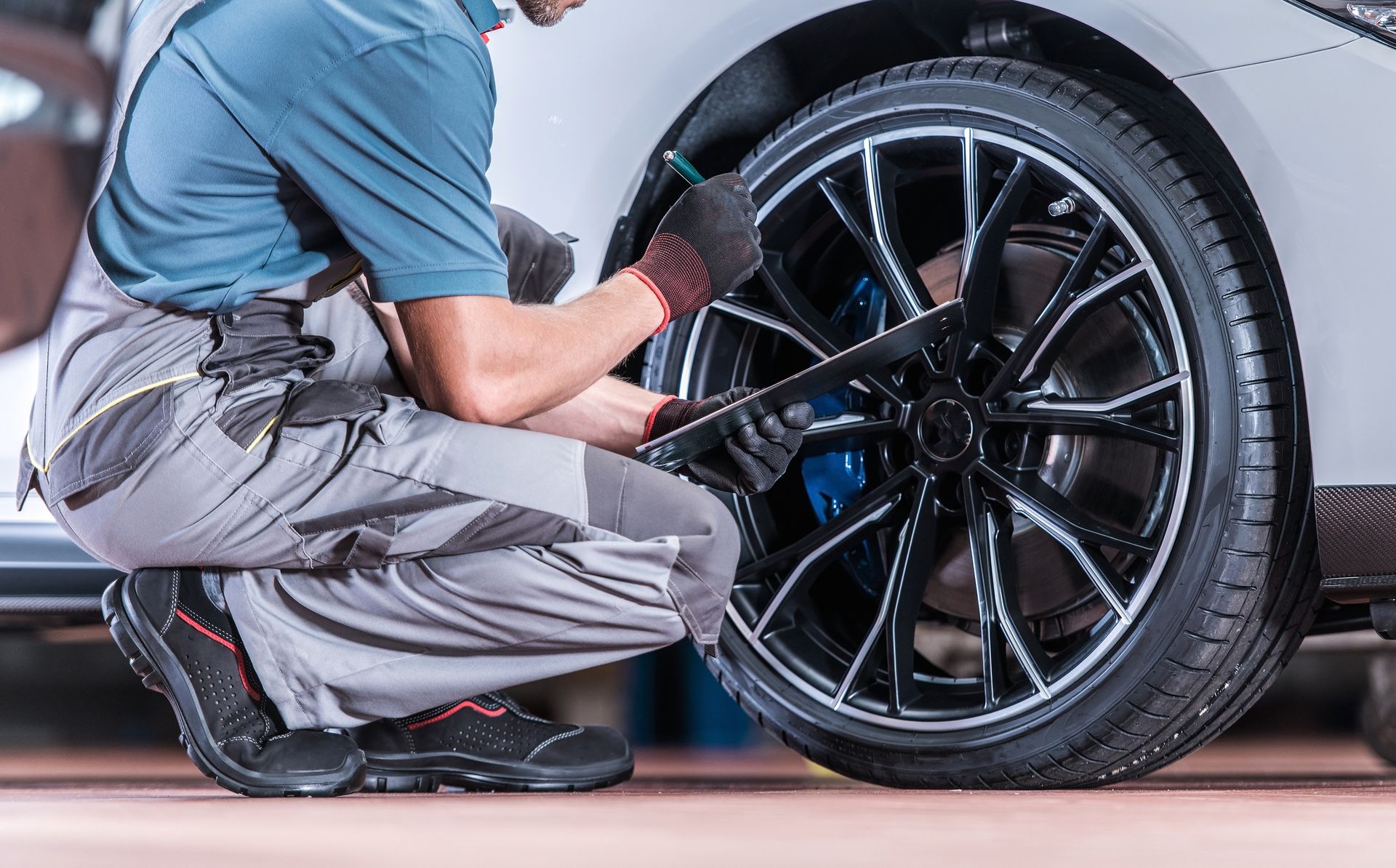 Mechanic kneeling beside a silver car, cleaning the black alloy wheel with a cloth.