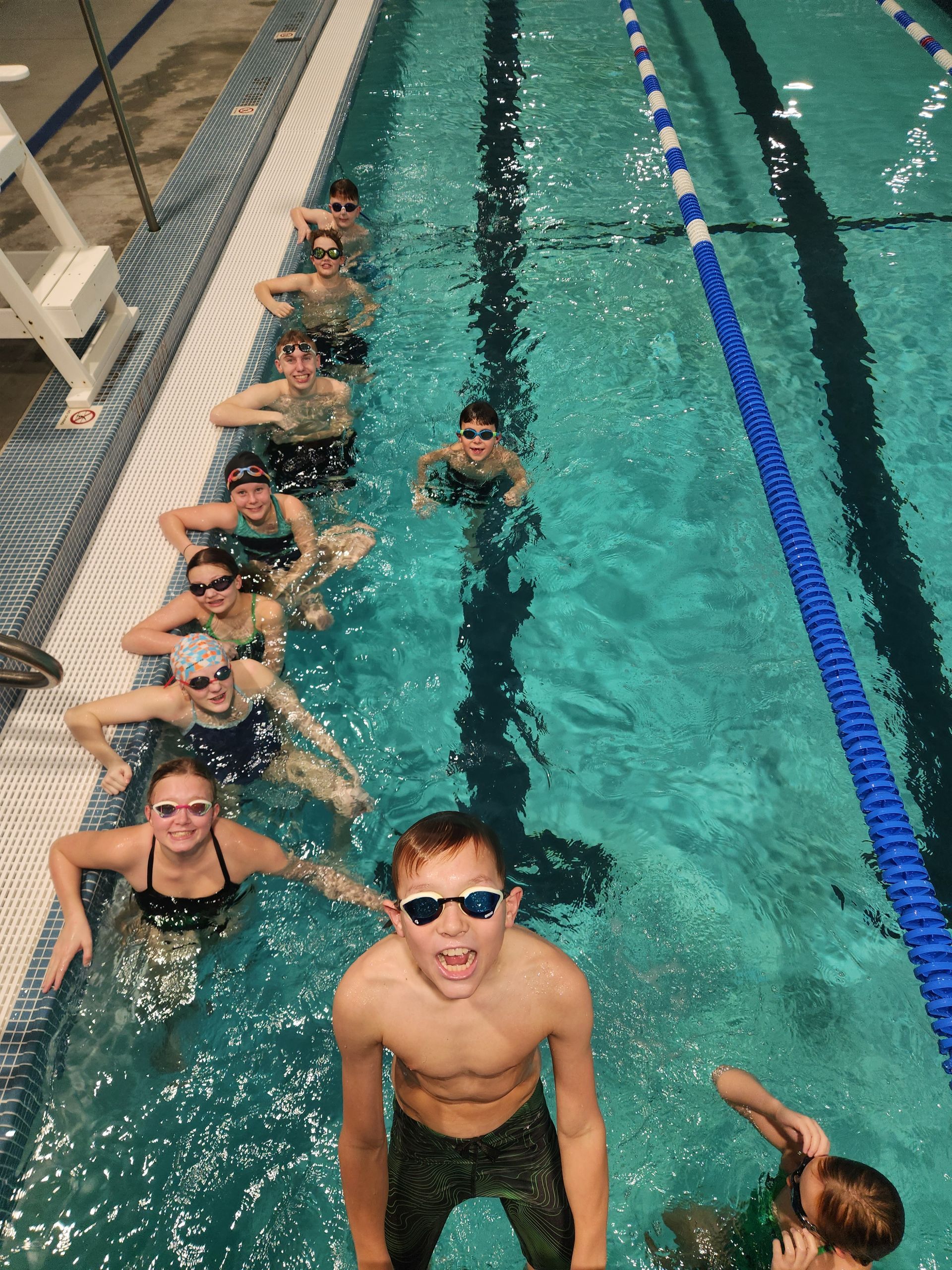Swimmers in a blue pool, some smiling, lined up near a lane marker.