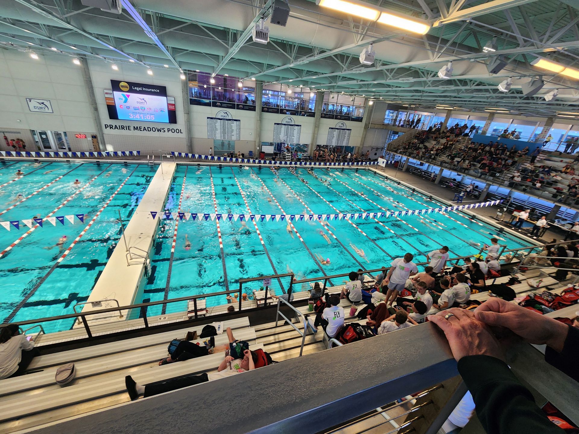 Indoor swimming pool, swimmers competing, spectators in stands, blue water, white lanes, bright lighting.