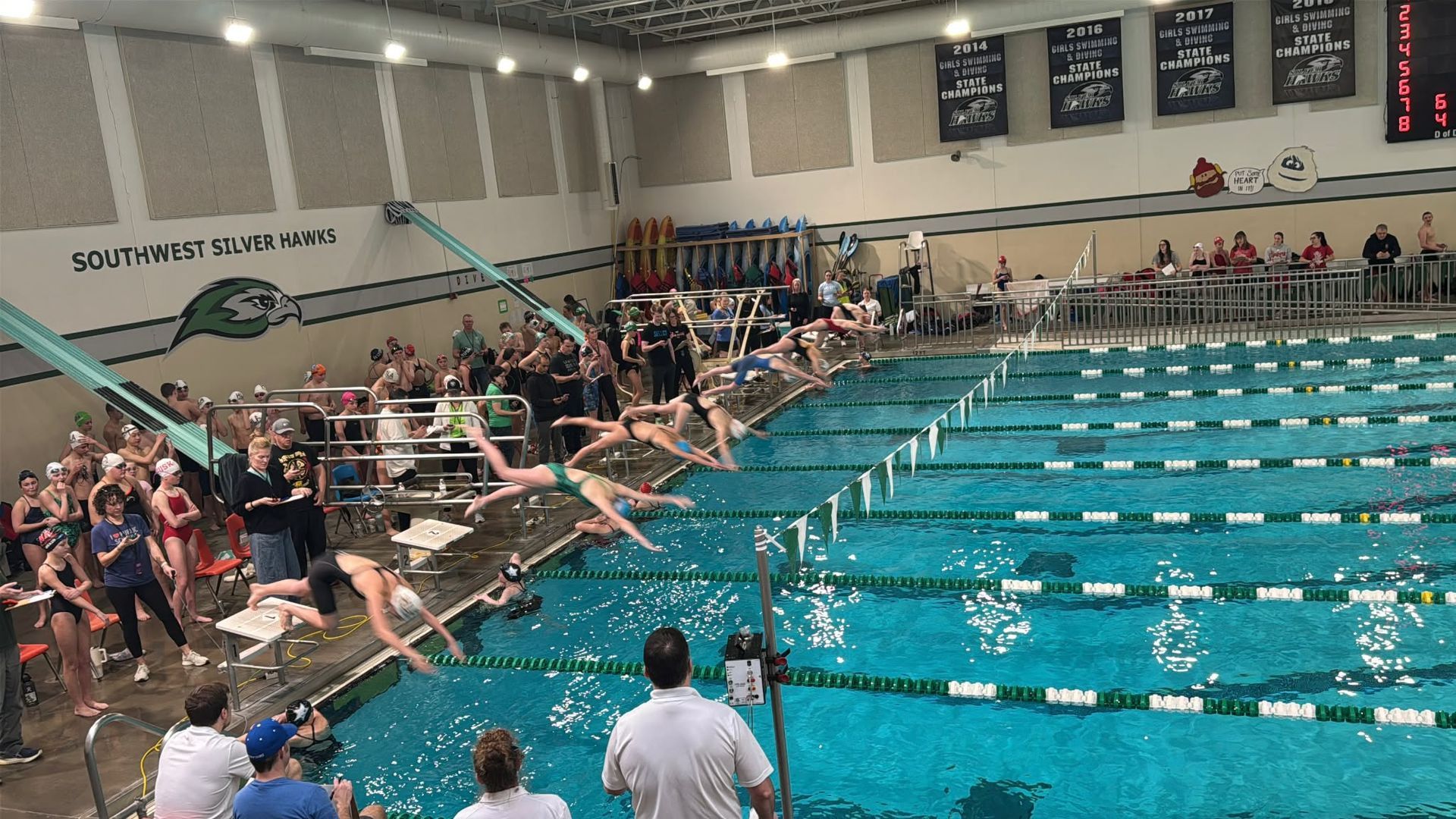 Swimmers diving into pool at a competition; spectators and coaches watch.