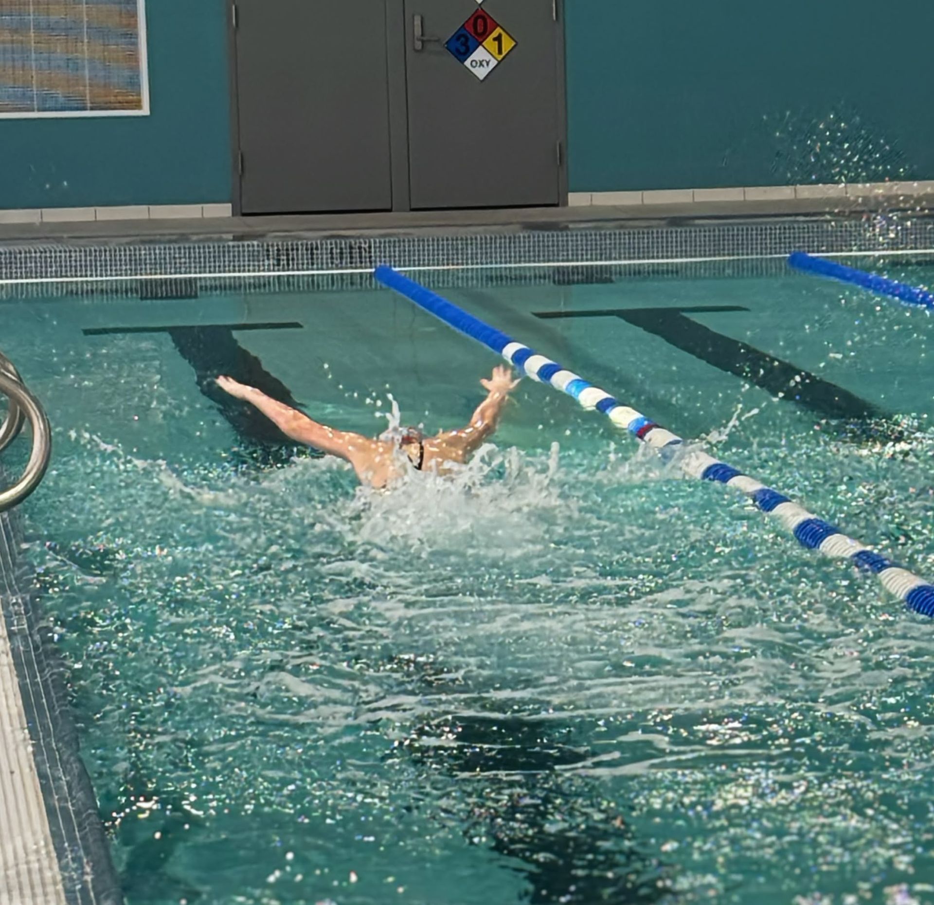 Swimmer in pool doing butterfly stroke. Blue water, splashes, lane markers. Building in background.