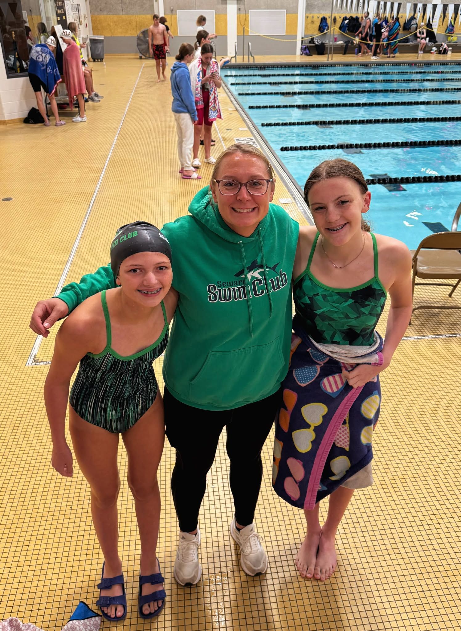 Three people pose by a pool. One in a green hoodie, two in swimsuits, smiling.