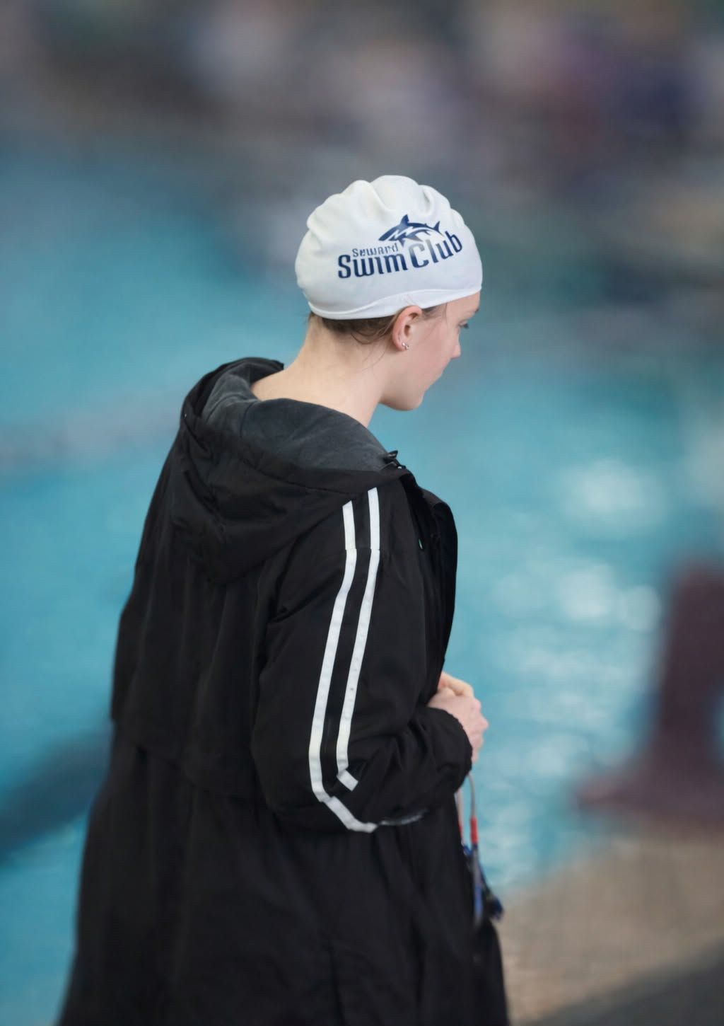 Swimmer wearing cap and black coat stands by pool.