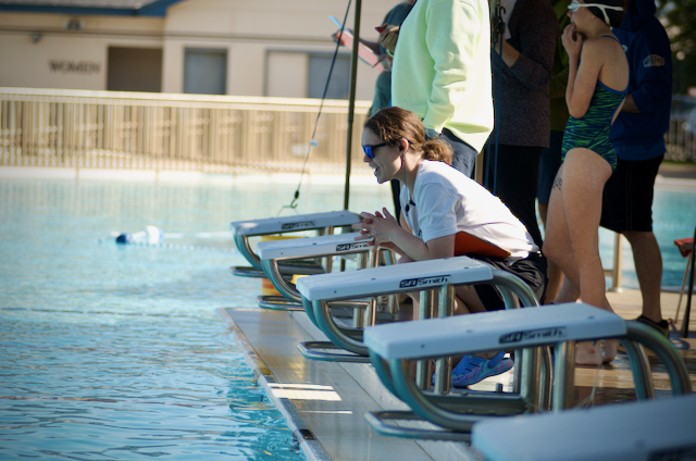 Swimmers at the edge of a pool, watching the competition. Woman in white shirt leaning on a block.