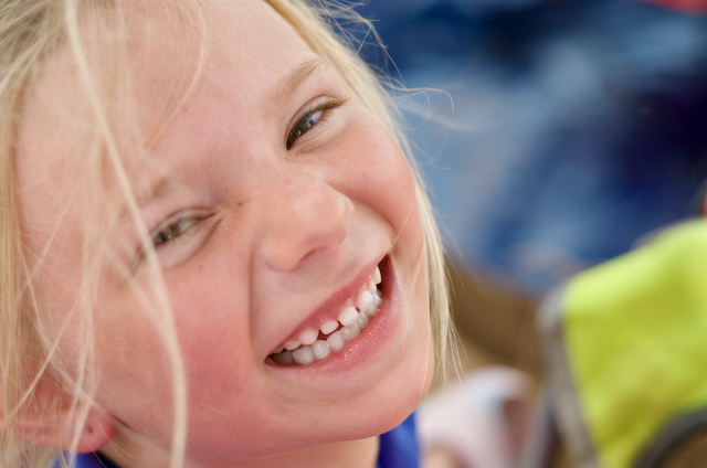 Blonde girl smiling mischievously, close-up shot. Partly obscured background, with a hint of blue and green.