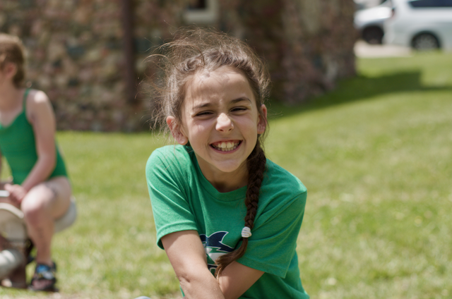 Girl with a braided brown ponytail smiles widely, wearing a green shirt, in a grassy outdoor setting.