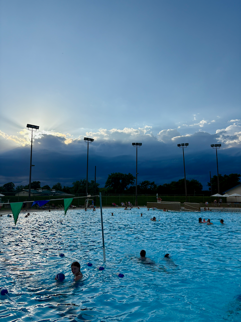 People swim in a large blue pool under a cloudy sky. Floodlights illuminate the scene.