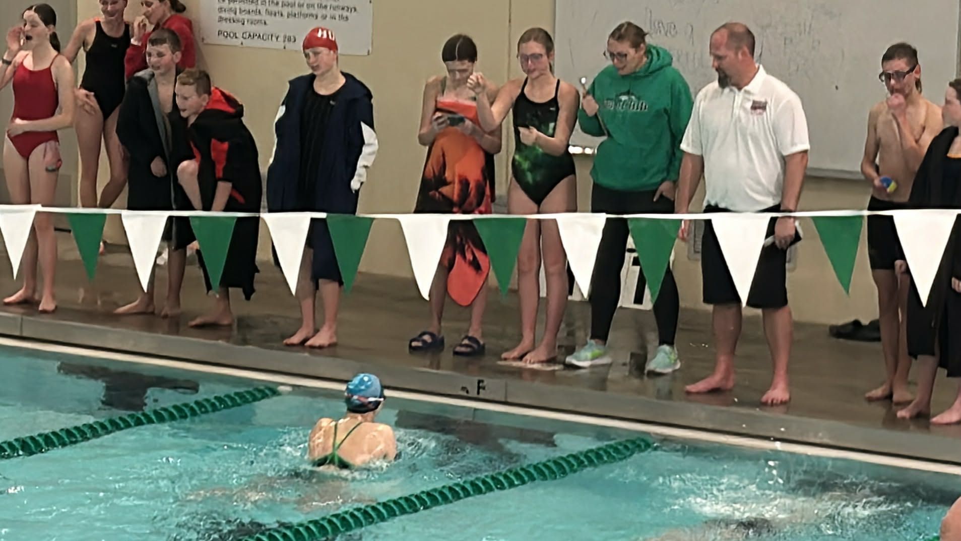 Swimmer in pool doing butterfly stroke; people watch from the side.