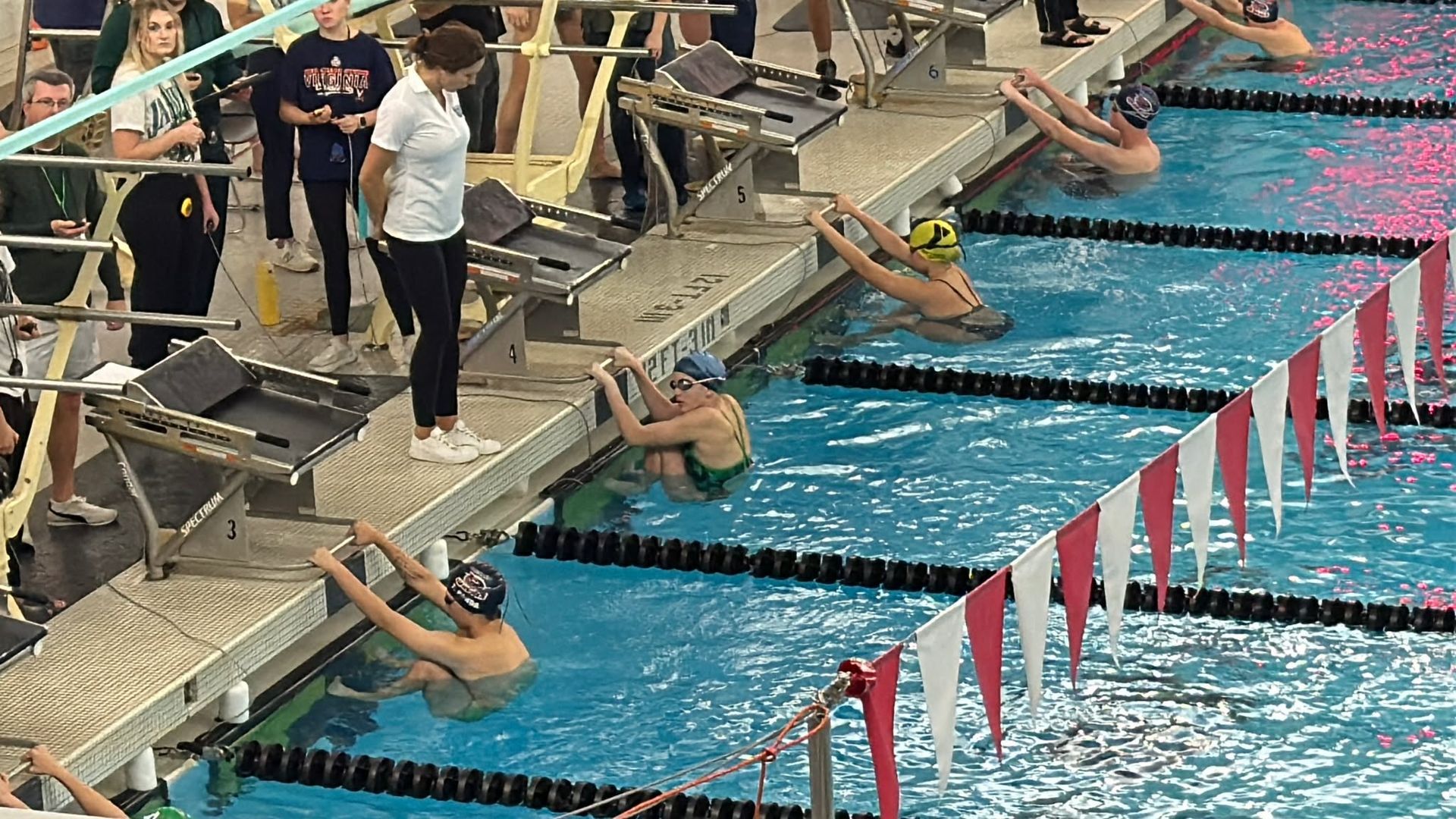 Swimmers in a pool preparing for a race, leaning on the edge with a coach nearby.