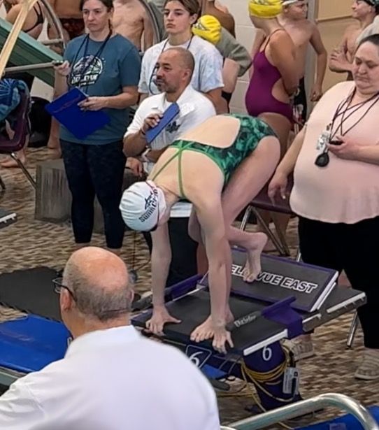 Swimmer in green swimsuit poised on a starting block, preparing to dive into a pool.