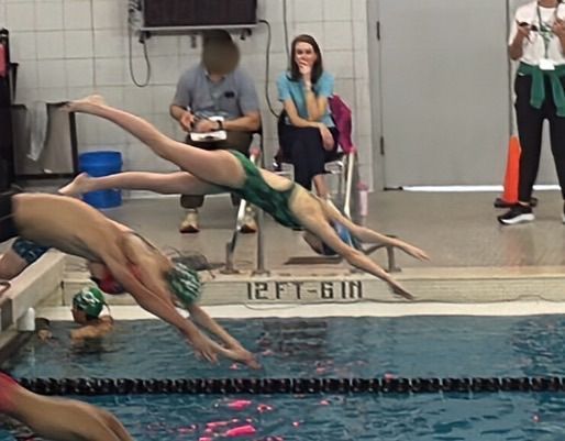 Swimmers diving into a pool; one in green swimsuit, others in caps, observers in background.