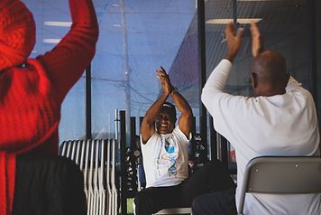 People seated indoors with arms raised, stretching or clapping in a group exercise class