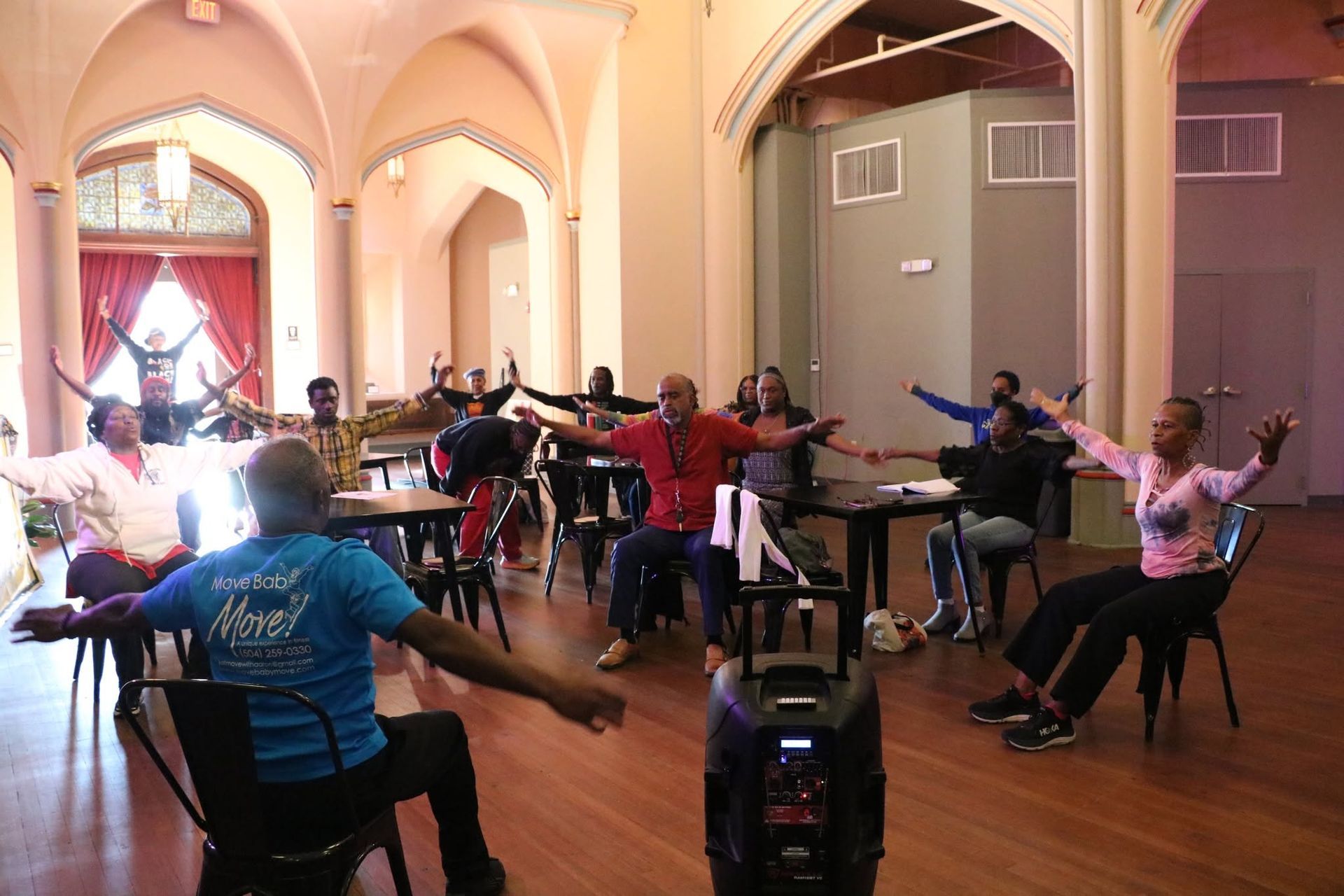 People seated in a bright hall doing arm stretches during a group exercise class