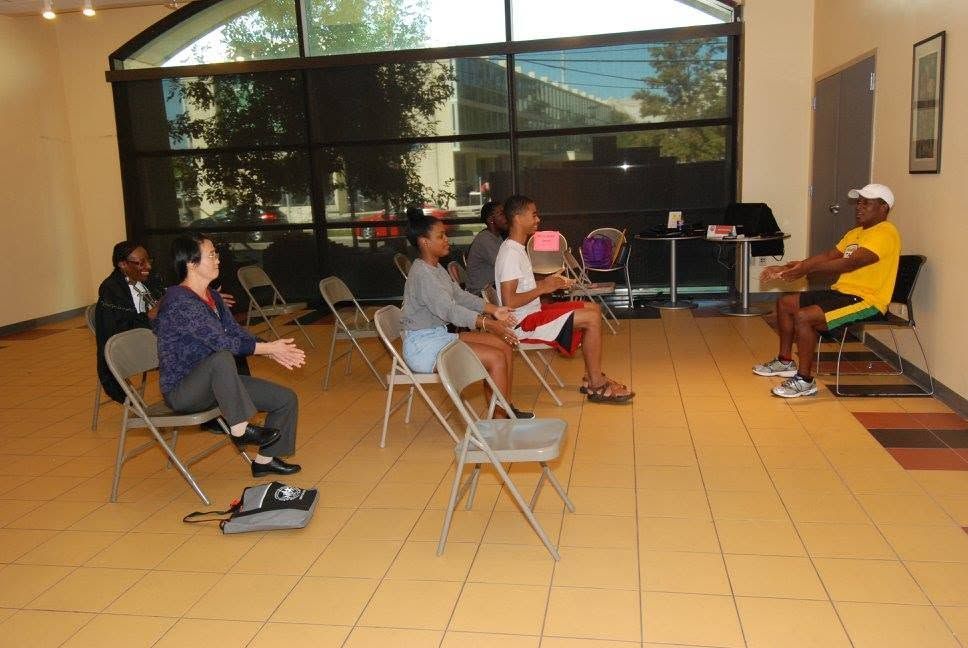 People seated in a social gathering on folding chairs in a bright indoor room with large windows.