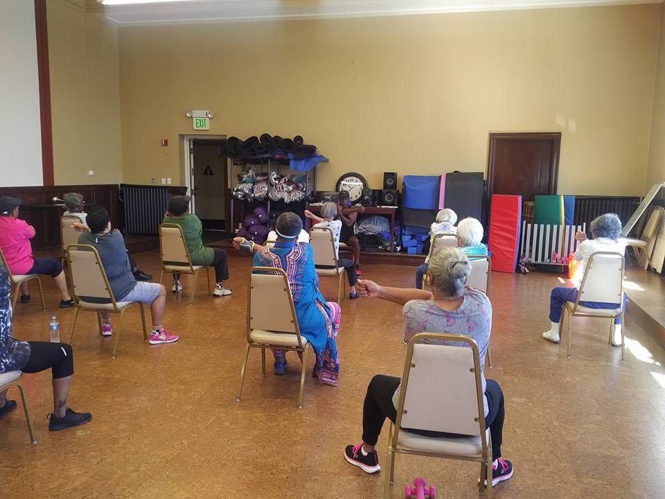 People seated on chairs in a gym-like room, facing a front stage or wall with colorful mats.
