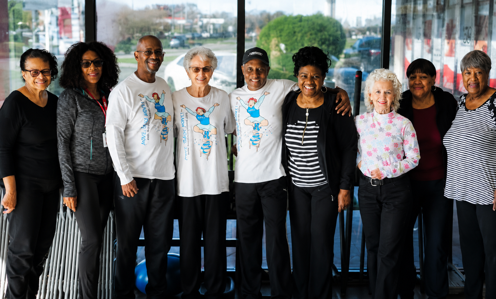 Eight people standing together indoors, wearing casual clothes in front of large windows.