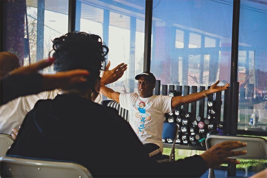 People seated in a bright room watching a person with arms outstretched leading a group activity
