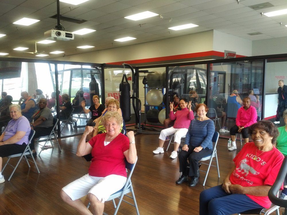 People seated in a bright fitness studio, following an exercise class with mirrors and windows.