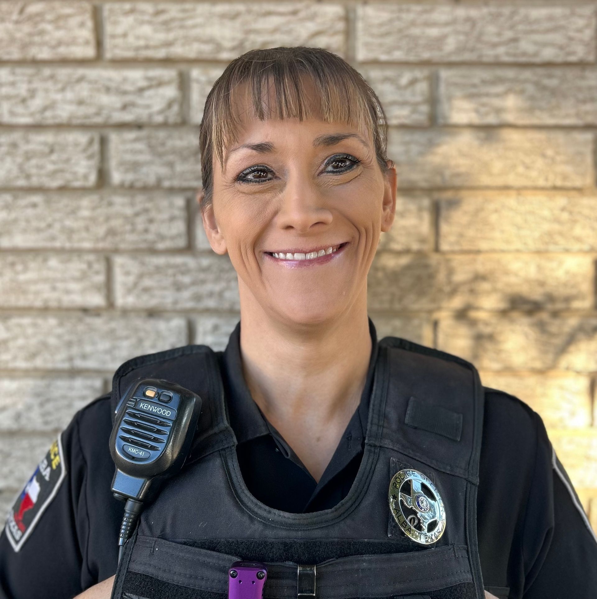 A woman in a police uniform is smiling and holding a walkie talkie.