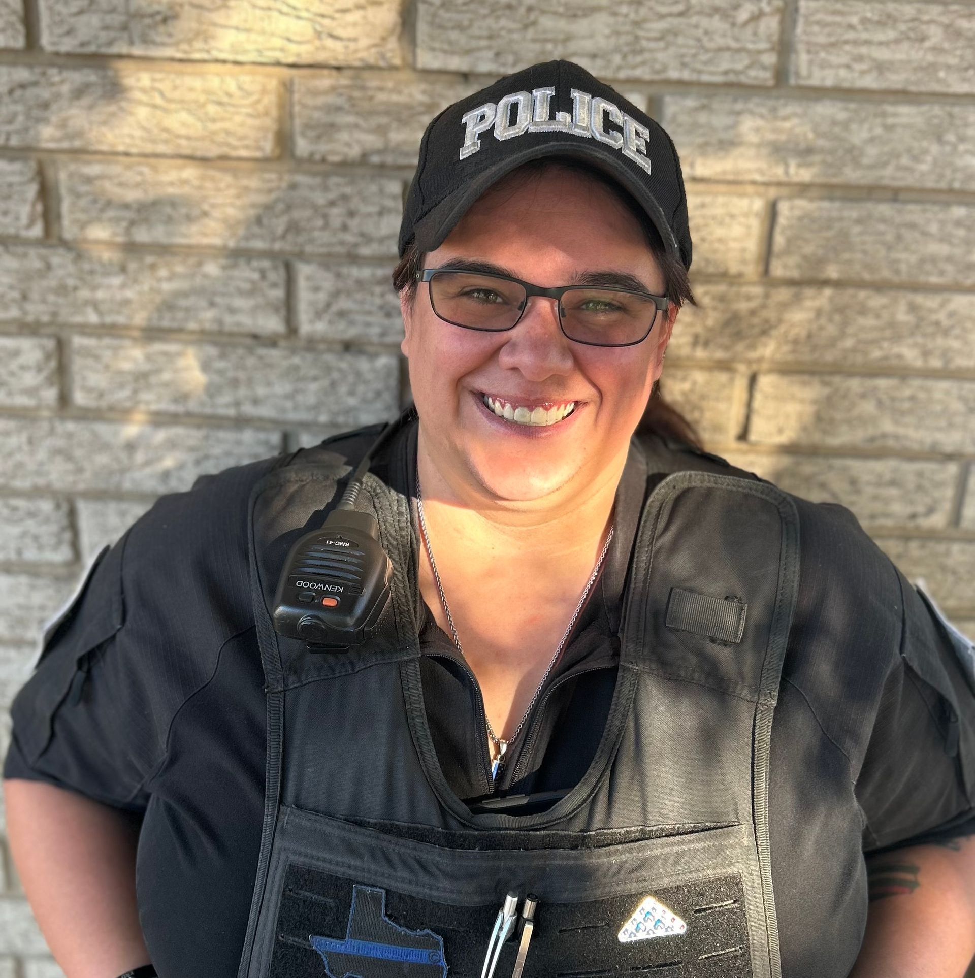 A woman wearing a police hat and vest is smiling in front of a brick wall.