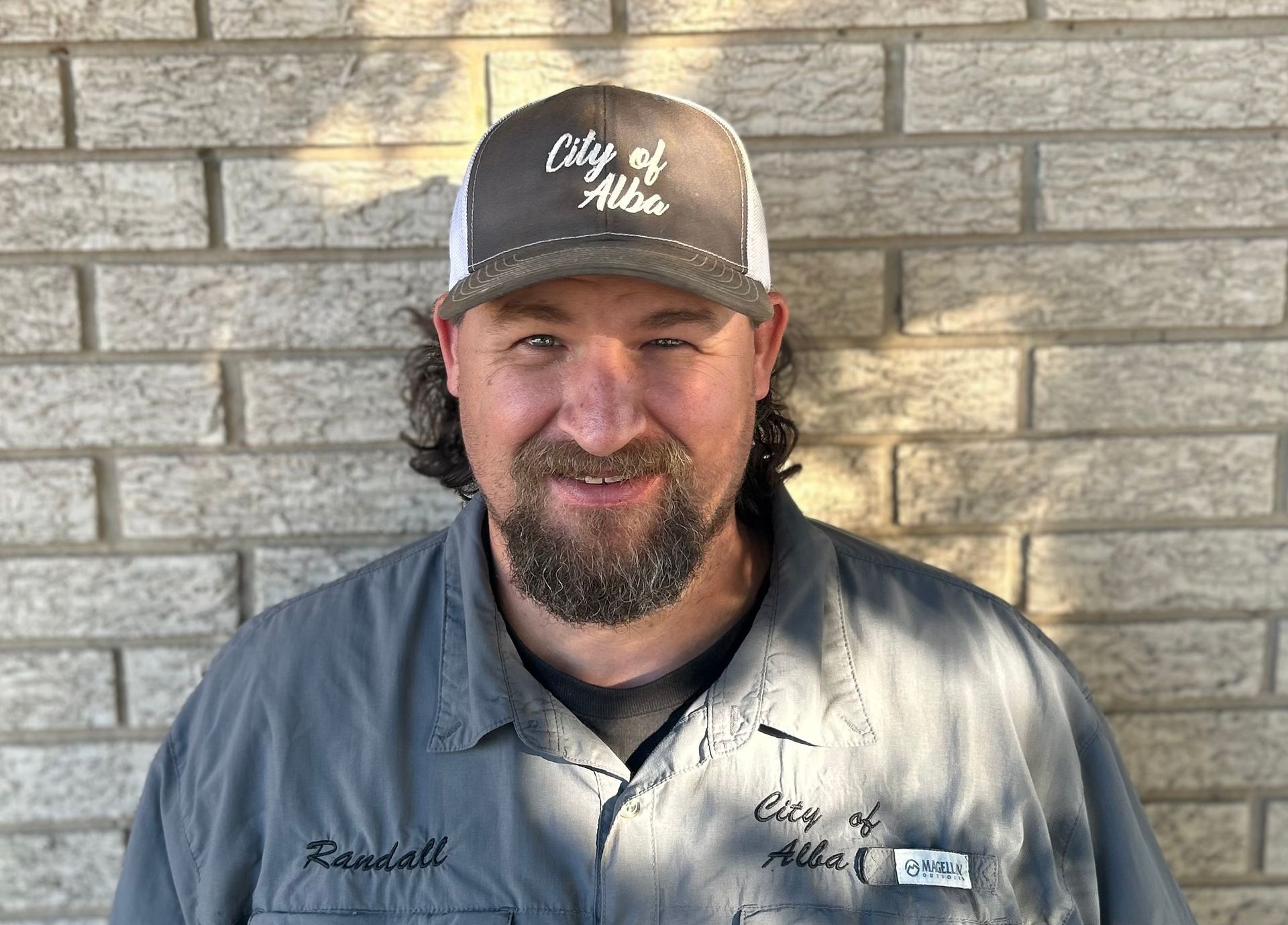 A man with a beard and hat is smiling in front of a brick wall.