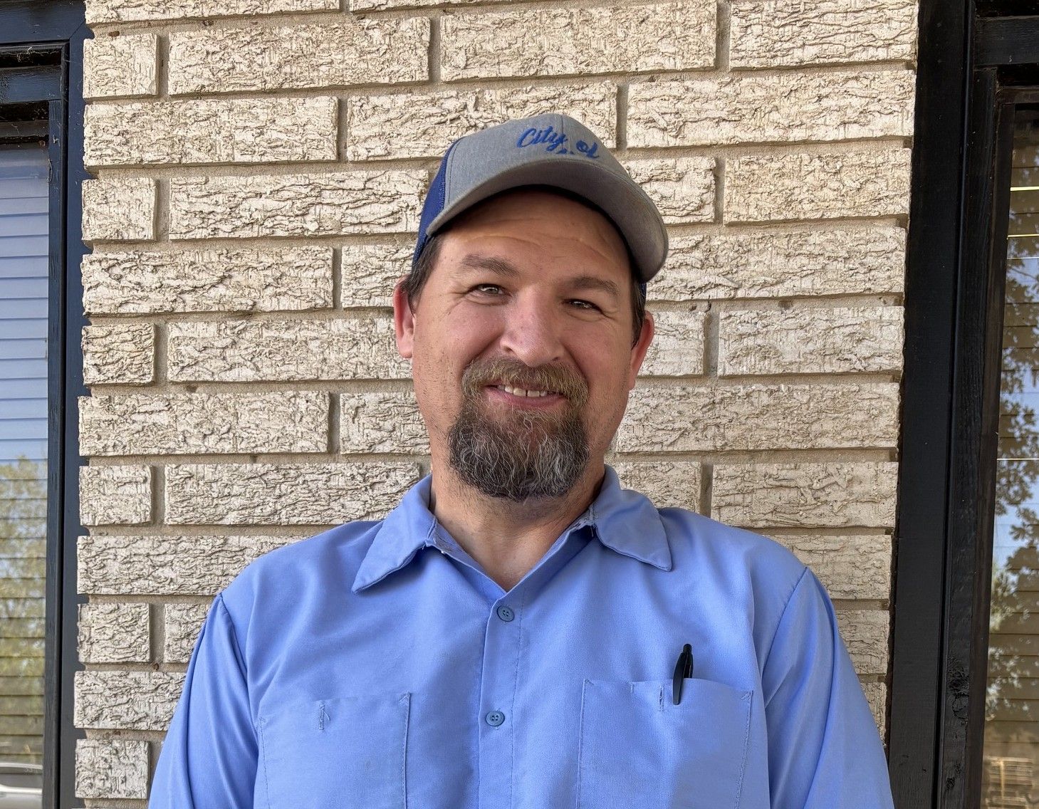 A man with a beard and hat is smiling in front of a brick wall.