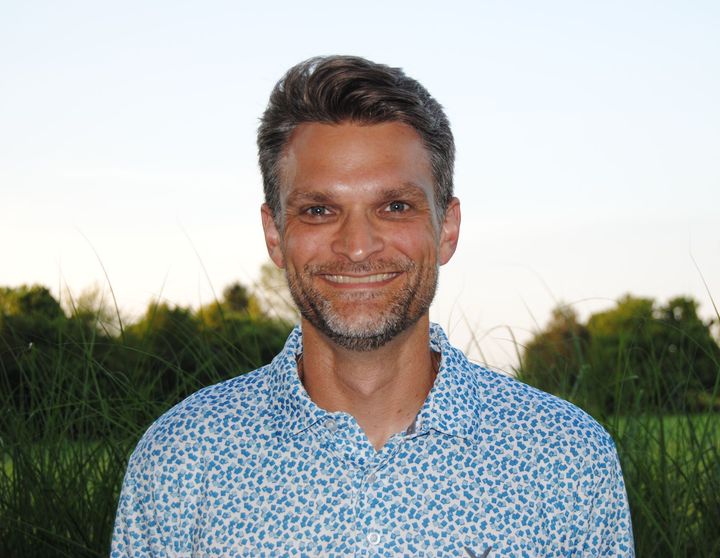 Smiling man in a blue patterned shirt standing outdoors with grass and a bright sky behind him