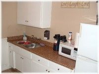 Small white kitchen with brown countertops, sink, cabinets, microwave, and refrigerator.