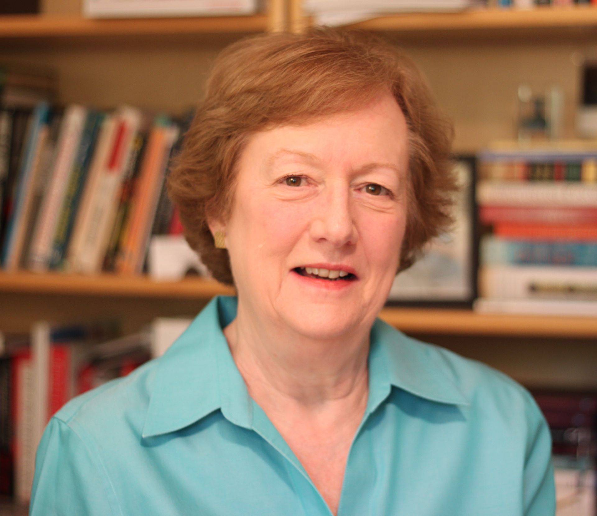 Photo of author Catherine Mathis sitting in front of bookshelves. Catherine has red hair and is wearing a tourquise colored blouse.