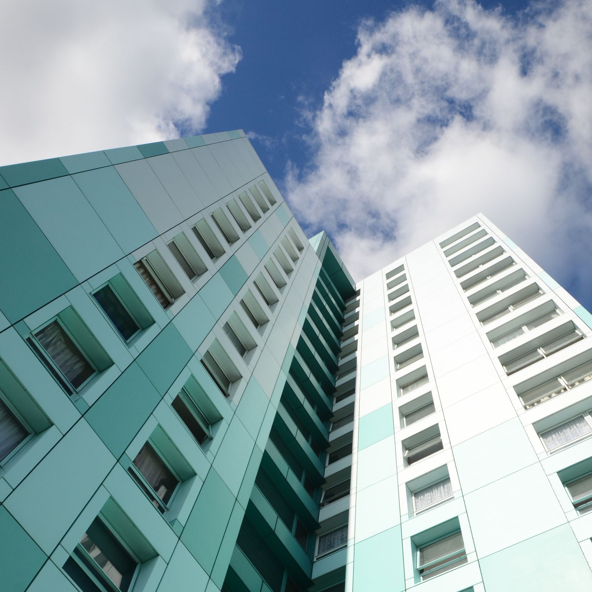 Low-angle view of two teal and white high-rise buildings against a blue sky with fluffy clouds.