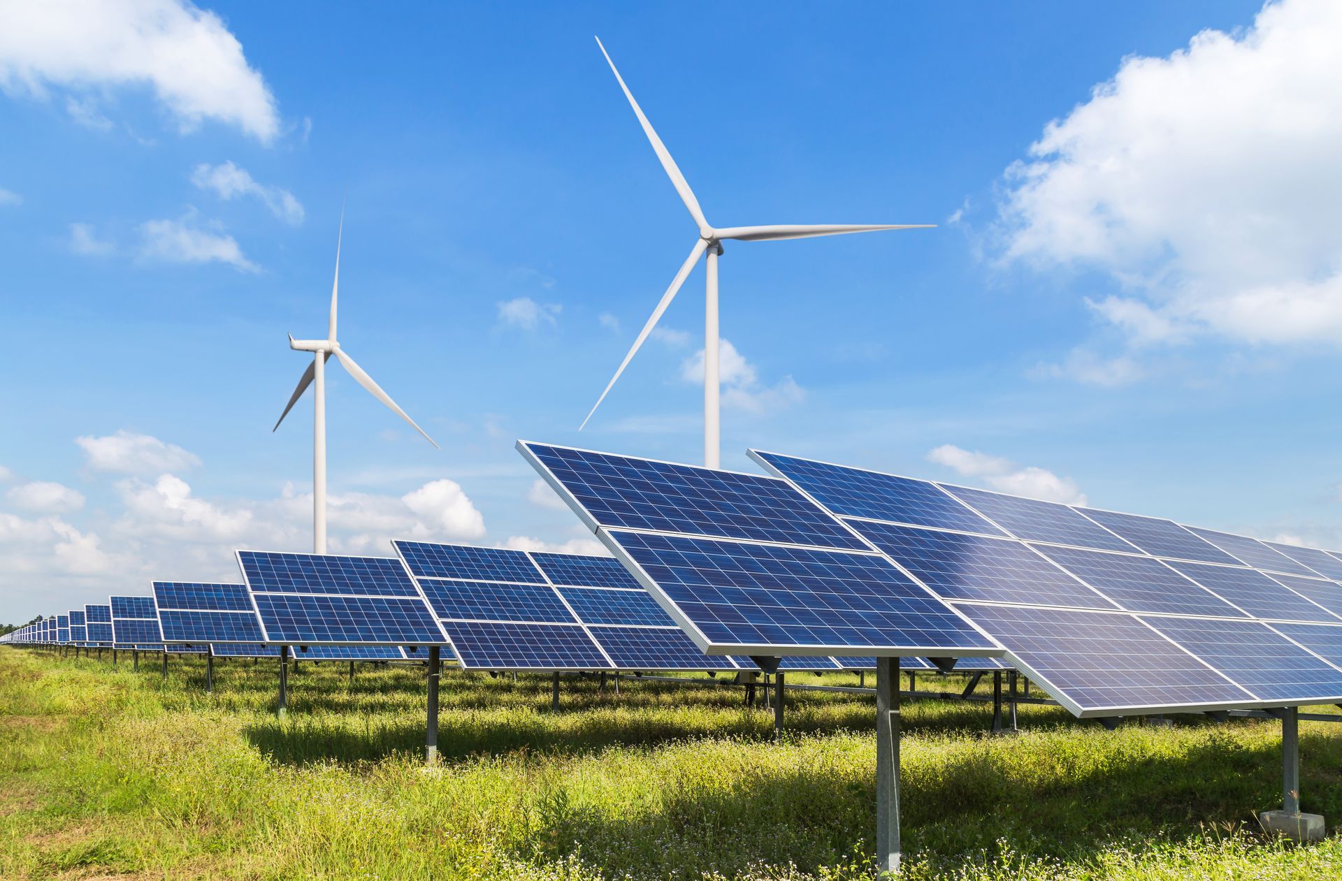 Solar panels and wind turbines in a grassy field under a blue sky.
