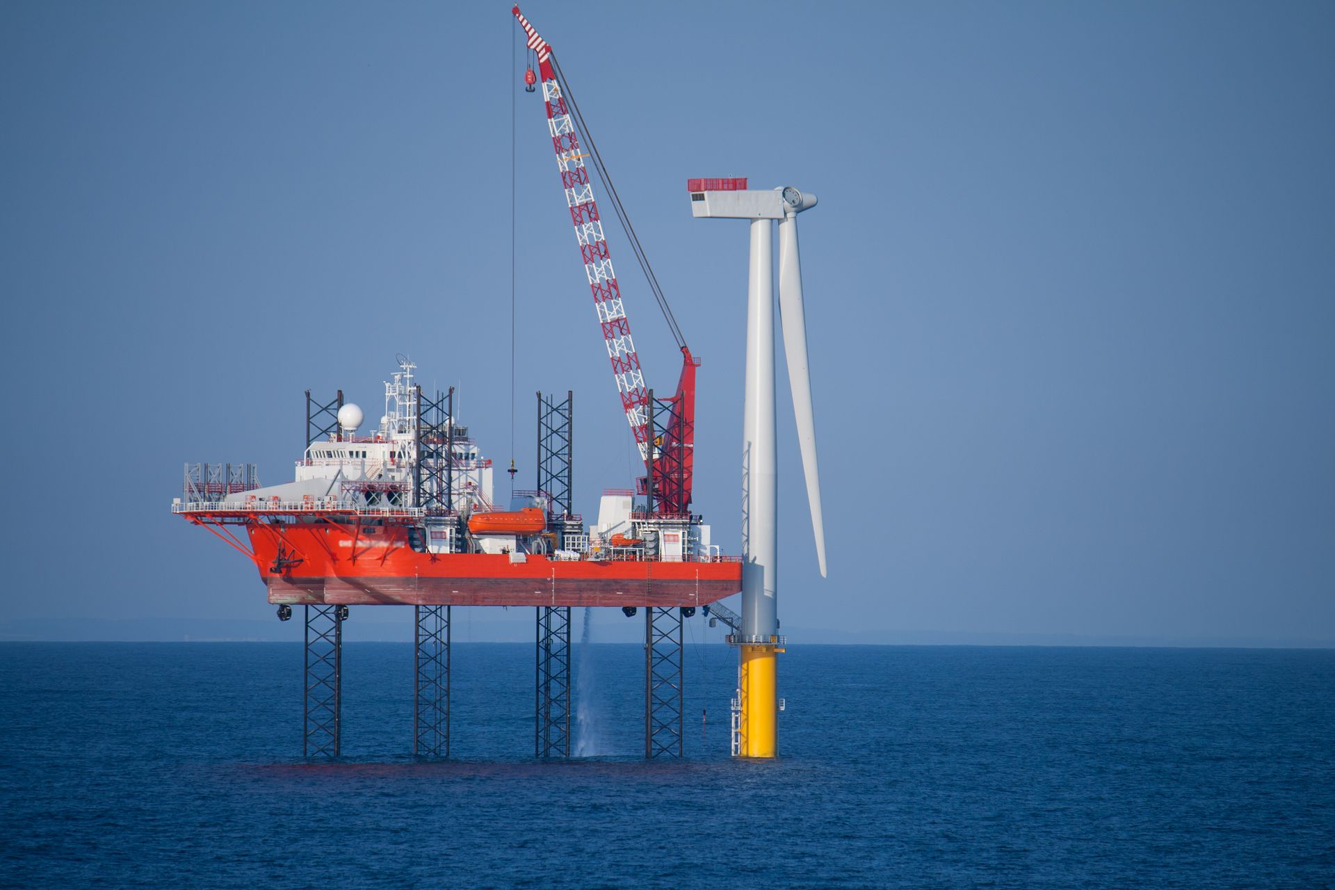 Offshore wind turbine construction: red jack-up vessel with crane, partially erected turbine in sea.