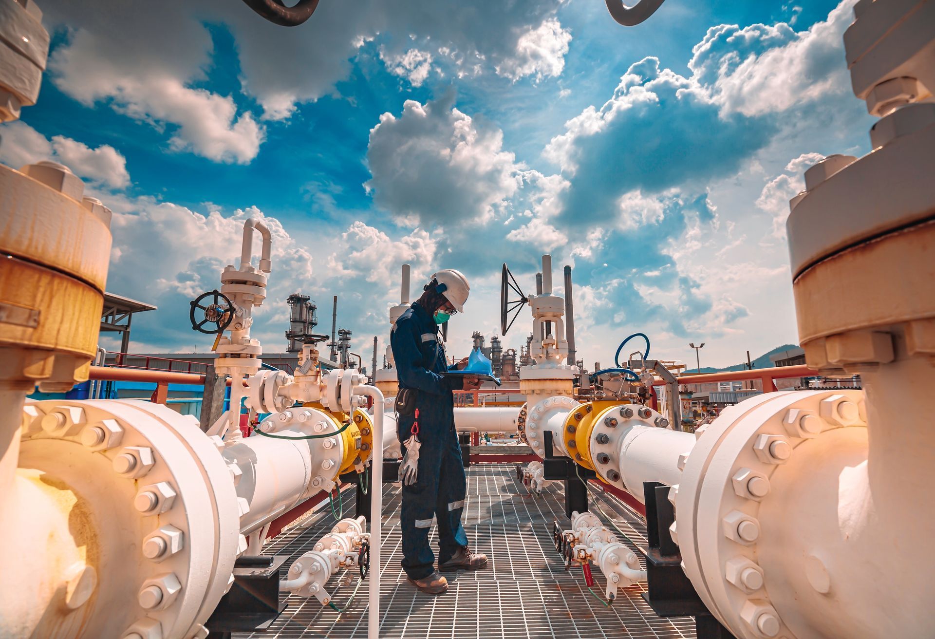 Technician inspecting pipeline equipment at an industrial facility; blue sky with clouds in the background.