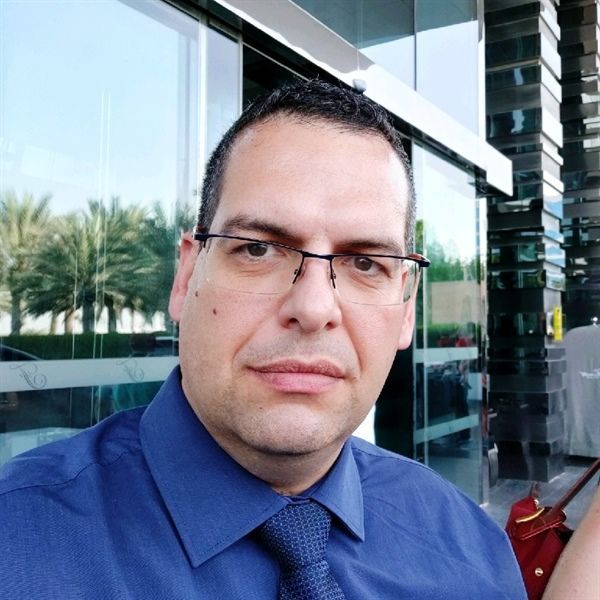 Man with glasses in blue shirt outdoors, looking at the camera.