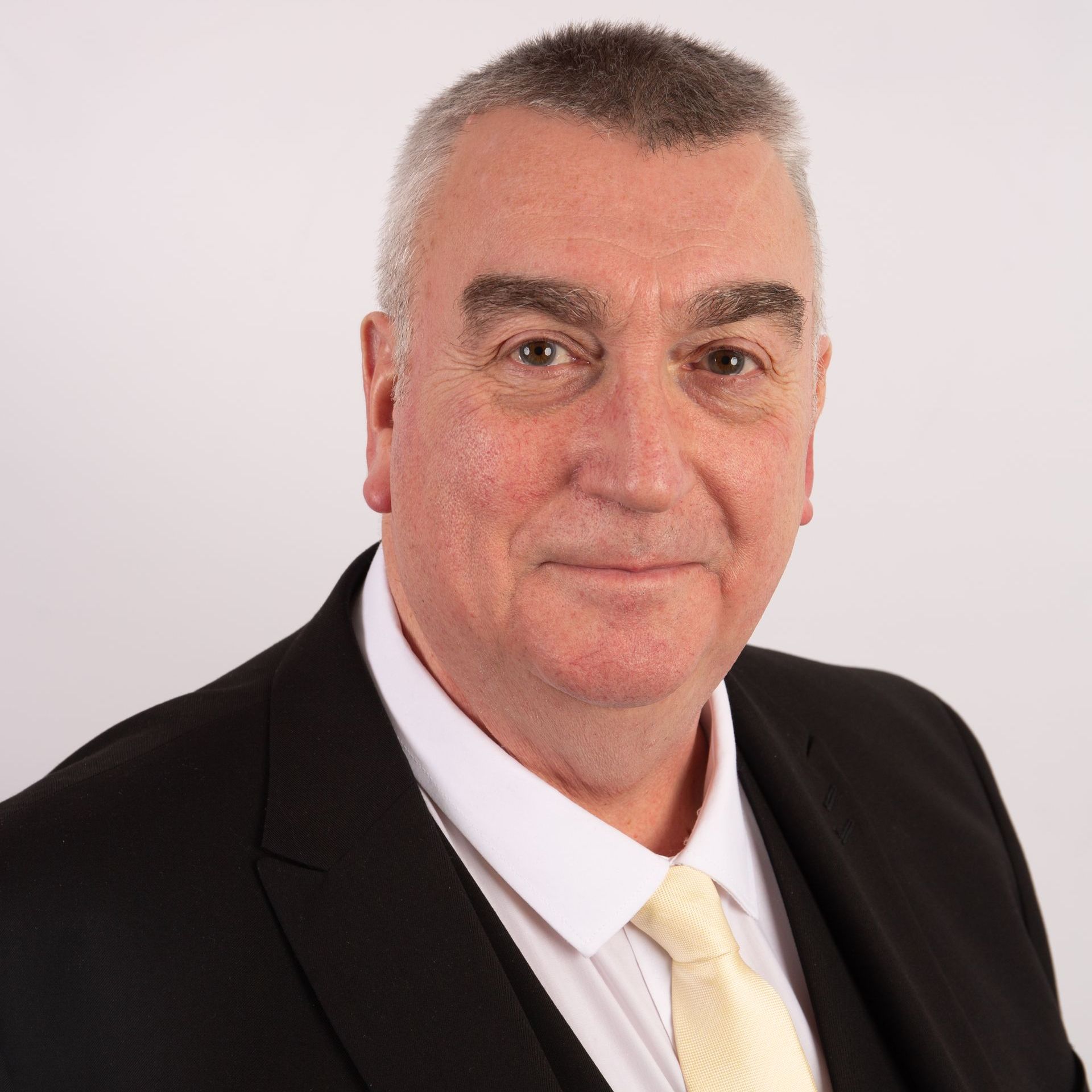 Man in a black suit with a cream tie, smiling against a white backdrop.