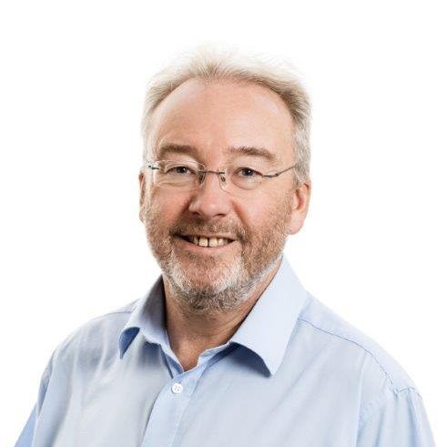 Man with glasses, graying hair, beard, smiling, wearing a blue collared shirt.
