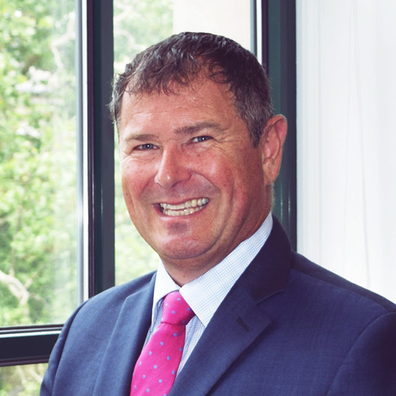 Man in suit smiling, pink tie, standing by window.
