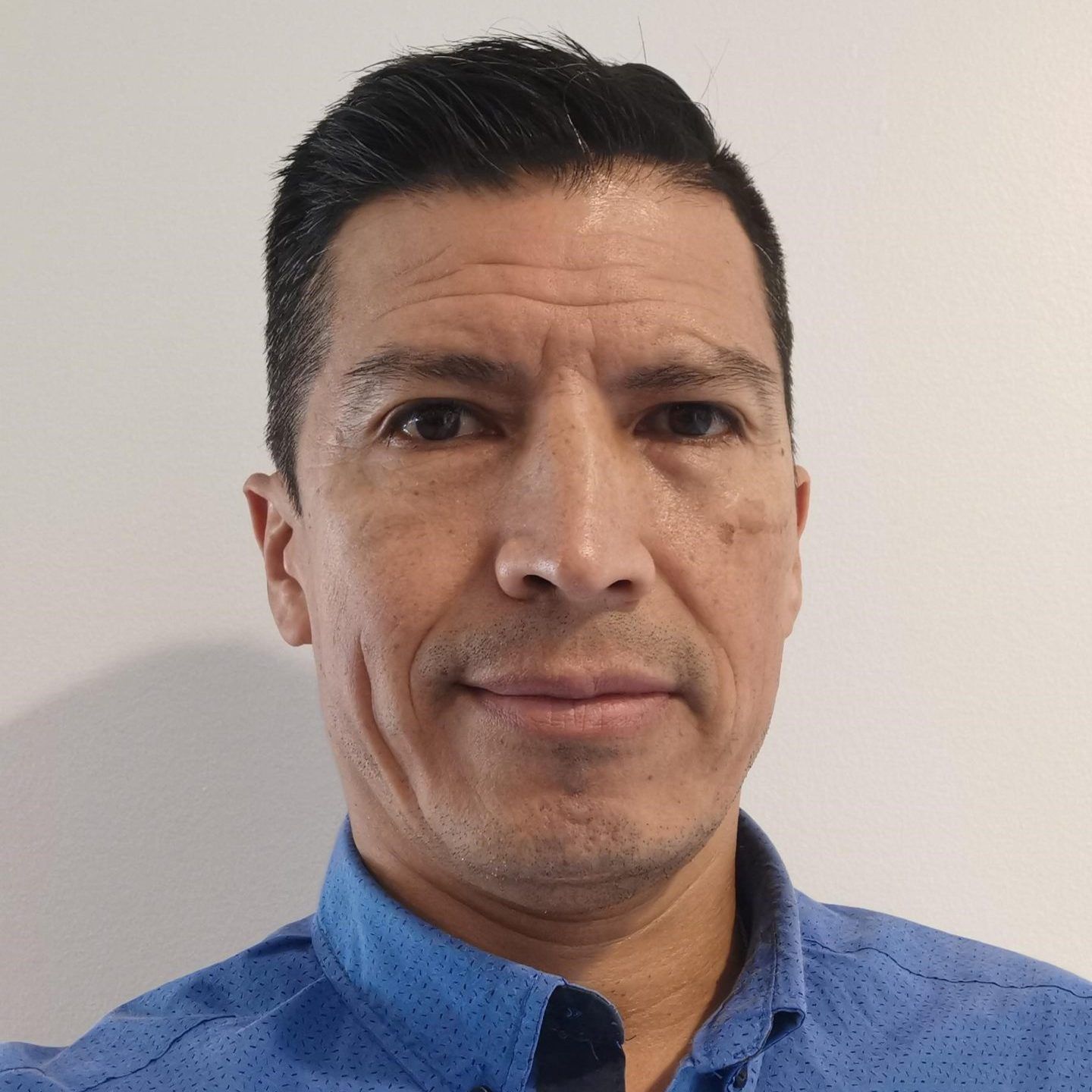 Man with short dark hair, wearing a blue shirt, looking at the camera. Neutral expression against a white wall.